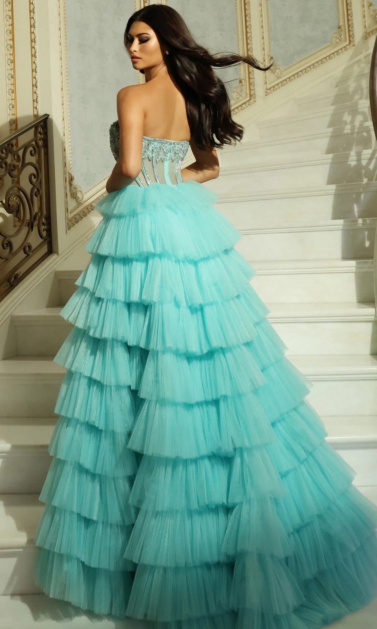 Wearing the Long Prom Dress: Jovani 44134, a woman in a strapless ruffle-tiered tulle gown stands on a marble staircase, facing away and looking over her shoulder.